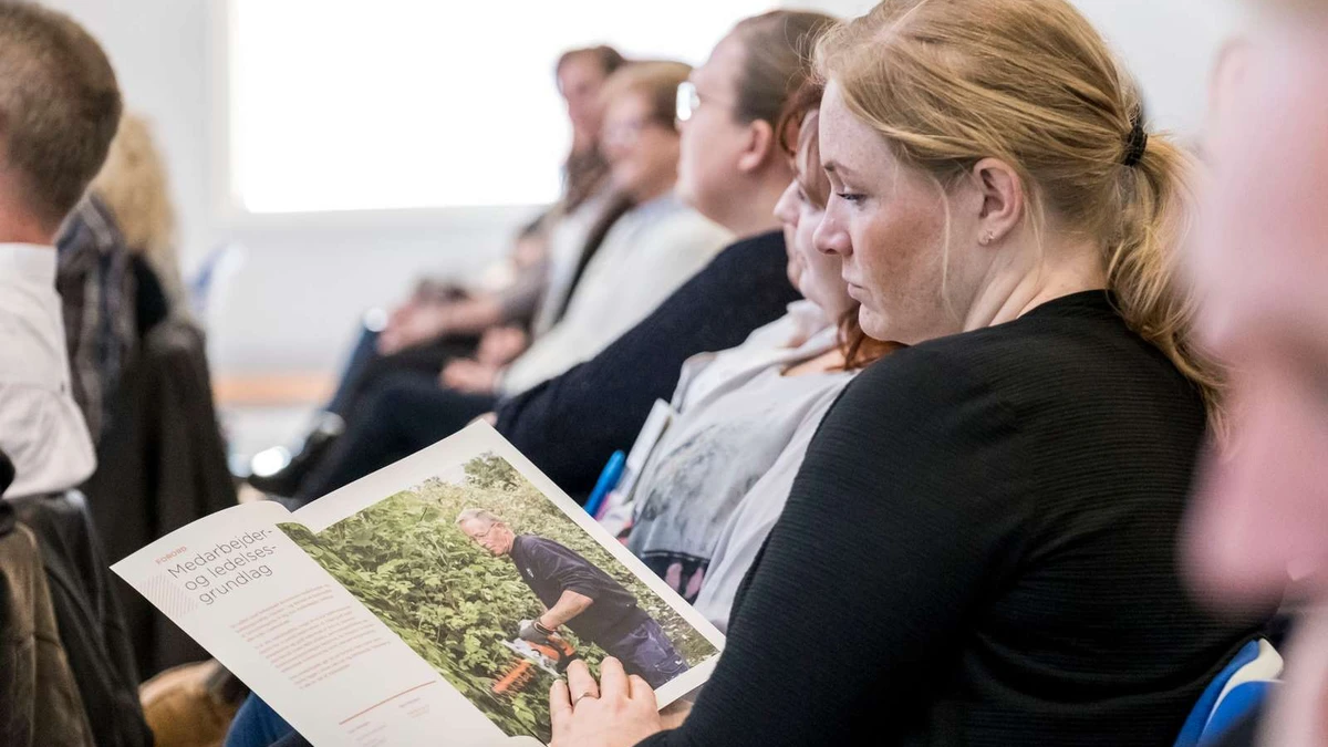 Kvinde læser en A-4 publikation fra stolerække med mange andre deltagere omkring hende