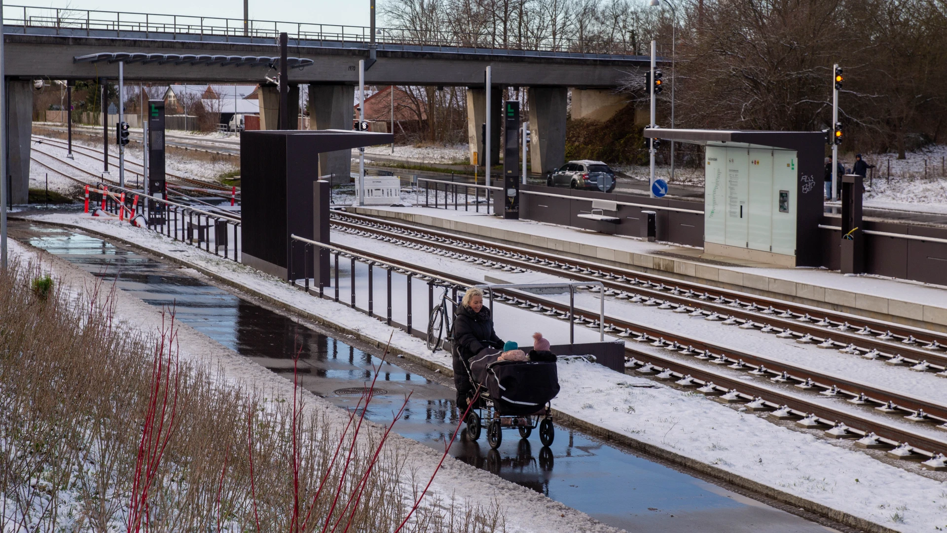 Kvinde går med barnevogn foran letbanesporet og med en jernbanebro i baggrunden. Der er sne.