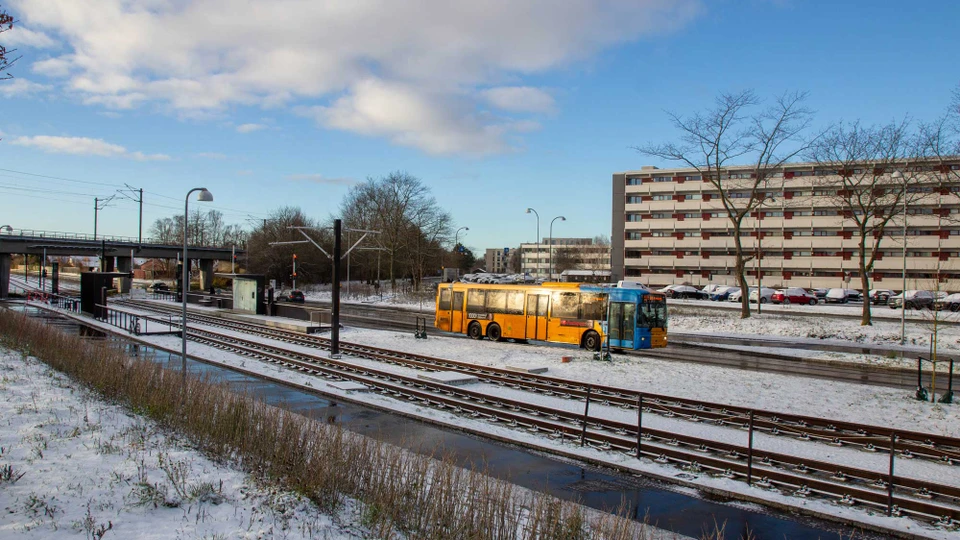 En gul bus med blå stribe kører ad en større vej. Der er letbaneskinner sideløbende med vejens forløb og i baggrunden ses en letbane-station.