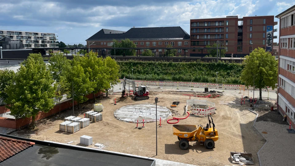 En grusbelagt plads midt i byrummet. Der står både minigraver, dumper og flere rammer fliser indenfor det afspærrede område.