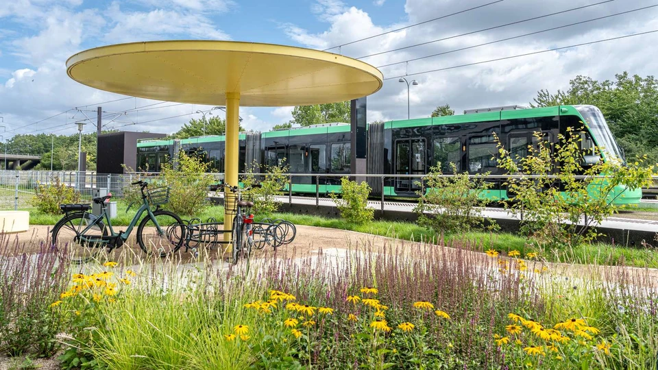 Grøn letbanetog ved en perron, i forgrunden en beplantet letbaneforplads med cykel i cykelstativ.