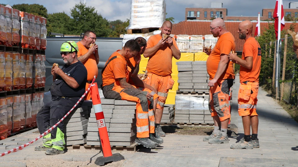 Håndværkere står og sidder på paller med fliser, imens de spiser flæskestegssandwiches