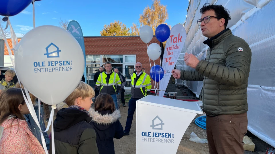 Mand taler og gør fagter foran et podie med balloner på hver side og børn og arbejdsmænd som publikum