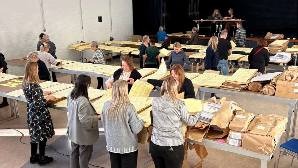 Tre lange borde med mange gule stemmesedler på og en masse fintællere ved bordene