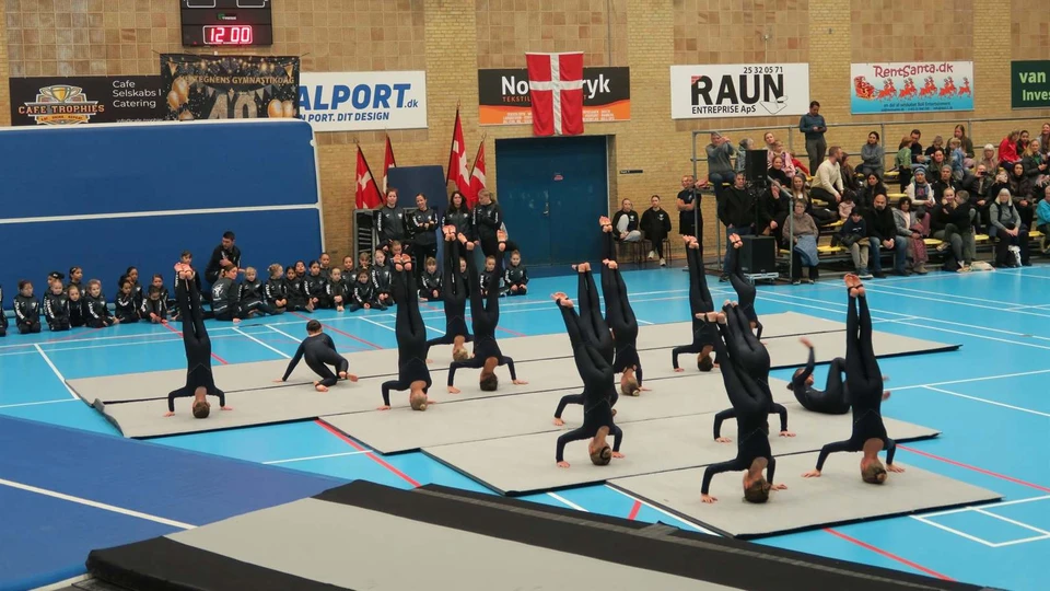 Omkring 12 unge gymnaster står på hoved på nogle måtter i en fyldt gymnastiksal