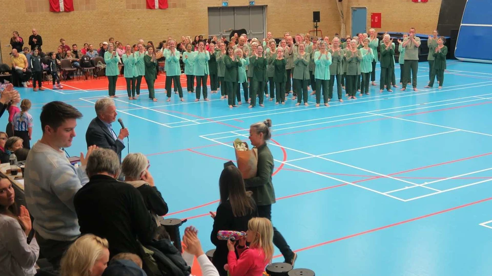 En kvinde med blomster i hånden går mod en mand med mikrofon. I baggrunden et stort gymnsatikhold der klapper.