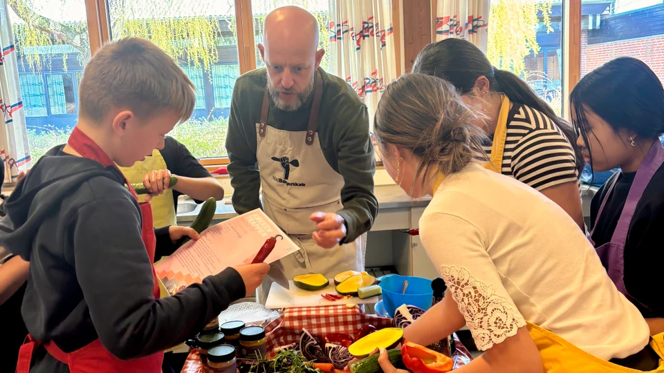 En mand med forklæde står foran fem større elever, der også bærer forklæde. På bordet imellem dem ligger mange madvarer.