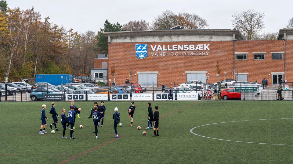 En gruppe børn står i cirkel på en fodboldbane og varmer op mellem spredte fodbolde