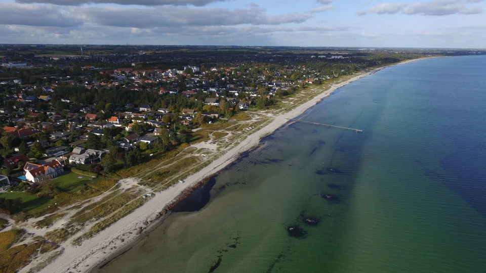Et dronebillede taget af en bugt, hvor der ud fra en sandstrand er en enkelt badebro. Inde på land ligger bebyggelsen relativt tæt på stranden.
