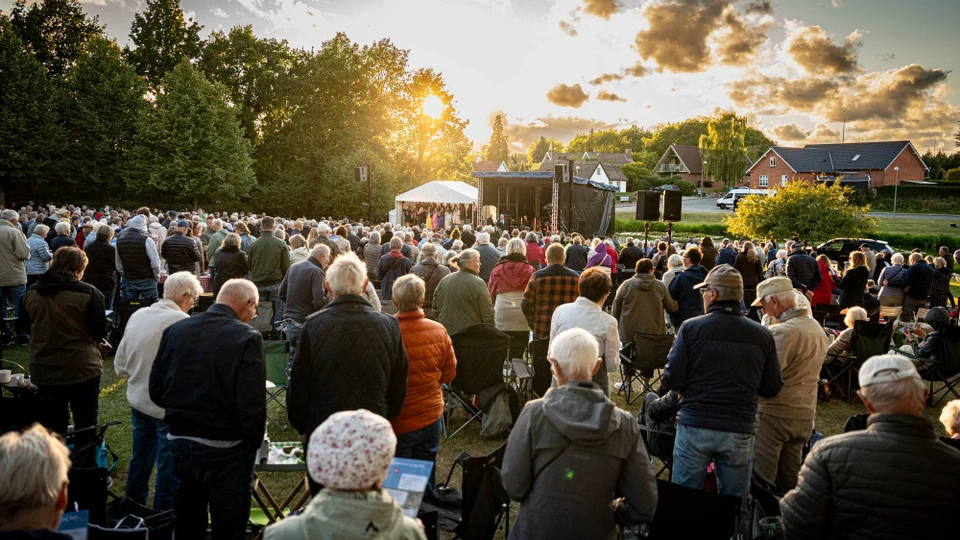 En koncertscene og foran dem er der fyldt ned mennesker på en smuk sommeraften.
