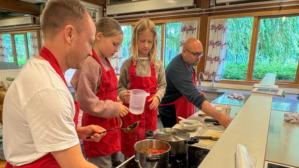 To piger står på skamler foran et komfur med to gryder på og to mænd på hver side af dem i køkkenet