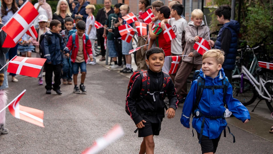 To drenge går og smiler til en masse børn, der står på række med flag og hepper på dem.