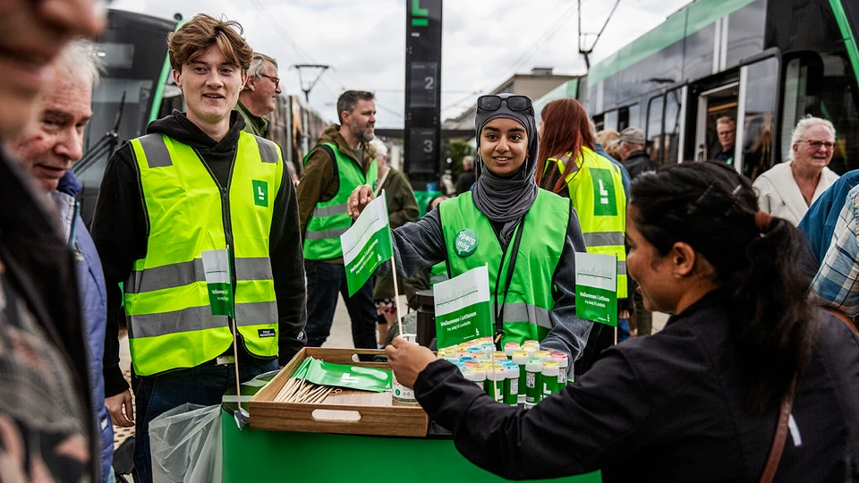 En tæt folkemængde ved en letbanestation. Der uddeles flag og sæbebobler af folk i veste.